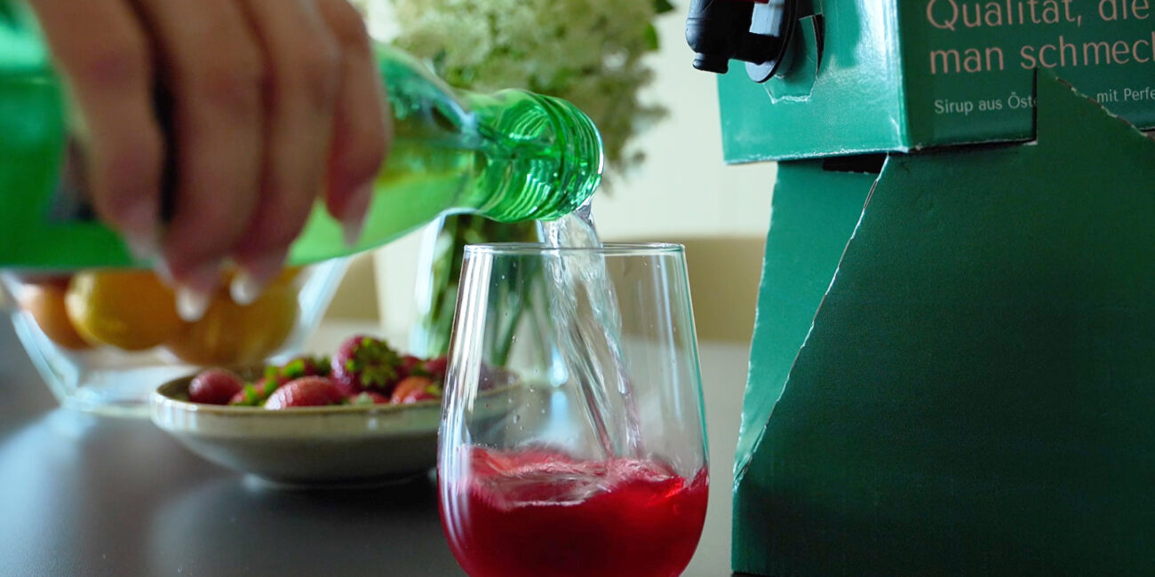 MySyrupBox und ein Glas auf einem Tisch wobei im Glas bereits Erdbeersirup ist und Sprudelwasser eingeschenkt wird.