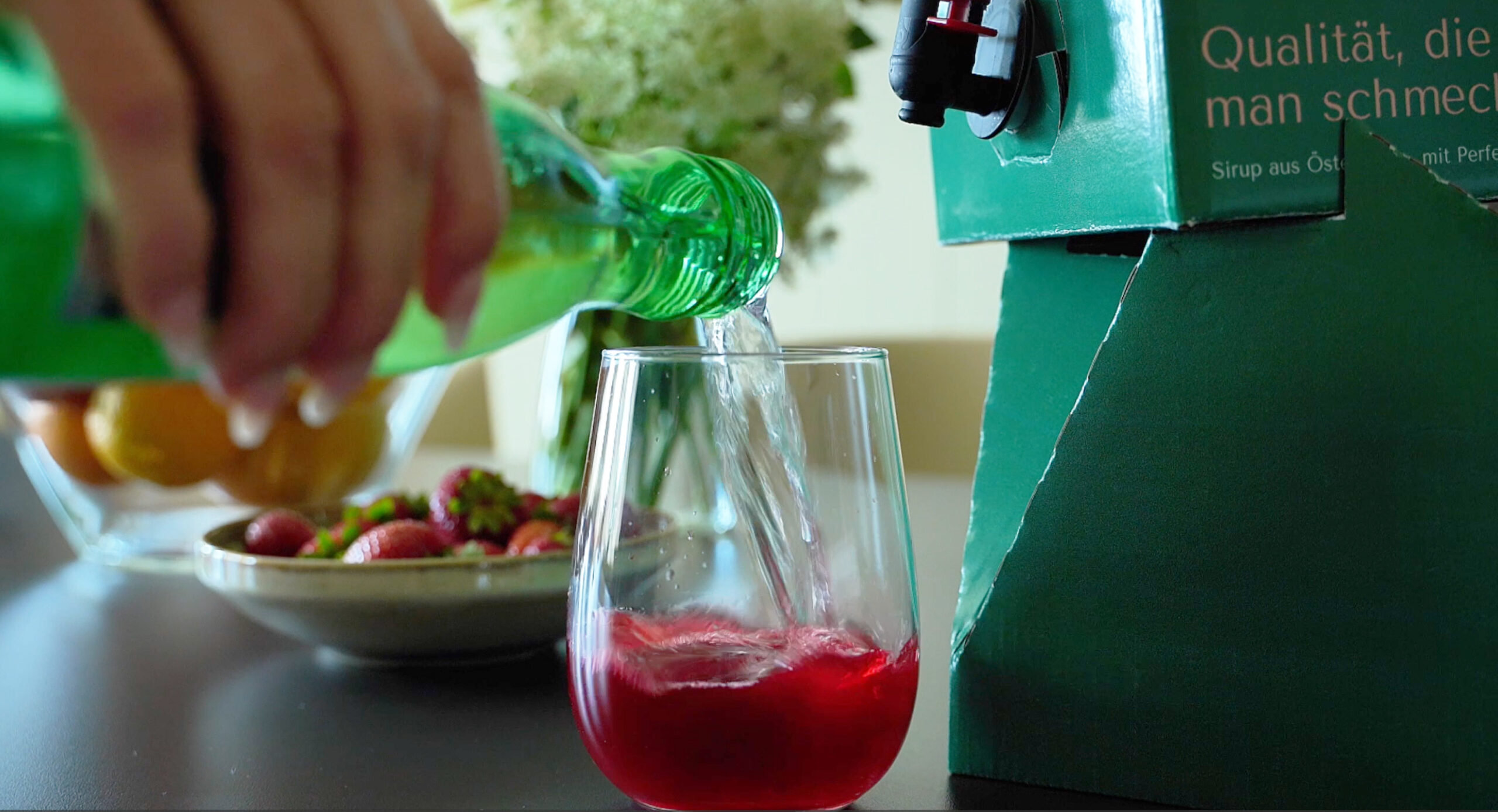 MySyrupBox und ein Glas auf einem Tisch wobei im Glas bereits Erdbeersirup ist und Sprudelwasser eingeschenkt wird.
